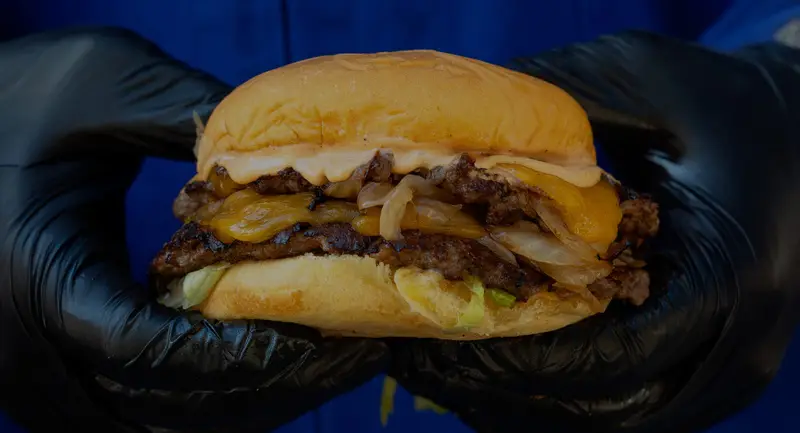 Hamburguesa doble con queso fundido sujeta con guantes negros sobre fondo azul.